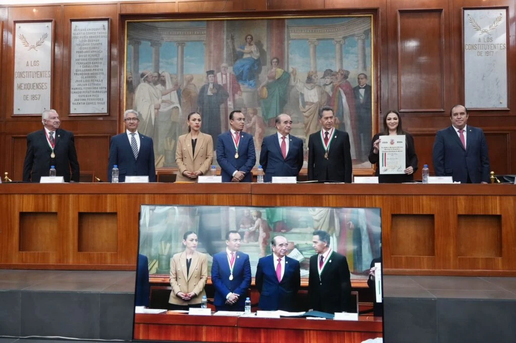Grupo de personas de pie detrás de una mesa en un acto protocolario; una de ellas sostiene un documento oficial mientras otras observan. En la parte inferior se ve una pantalla que proyecta la misma escena, y al fondo hay una pintura histórica y placas conmemorativas.