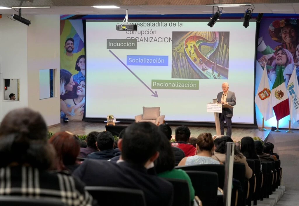 Auditorio con público sentado observando a una persona que expone en un pódium frente a una pantalla grande con una presentación sobre corrupción organizacional; se observan gráficos y texto proyectados, así como banderas institucionales a un costado.