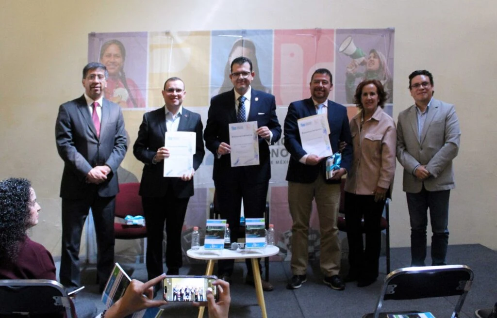 Seis personas de pie en un escenario sostienen documentos de reconocimiento durante un acto institucional; al frente se observa una mesa con libros y material informativo, mientras una persona del público toma una fotografía con un teléfono móvil.