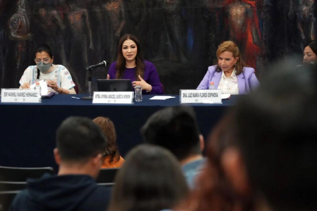 Panel de ponentes sentado detrás de una mesa con micrófono y gafetes, frente a una audiencia en un auditorio, con un mural artístico oscuro al fondo.