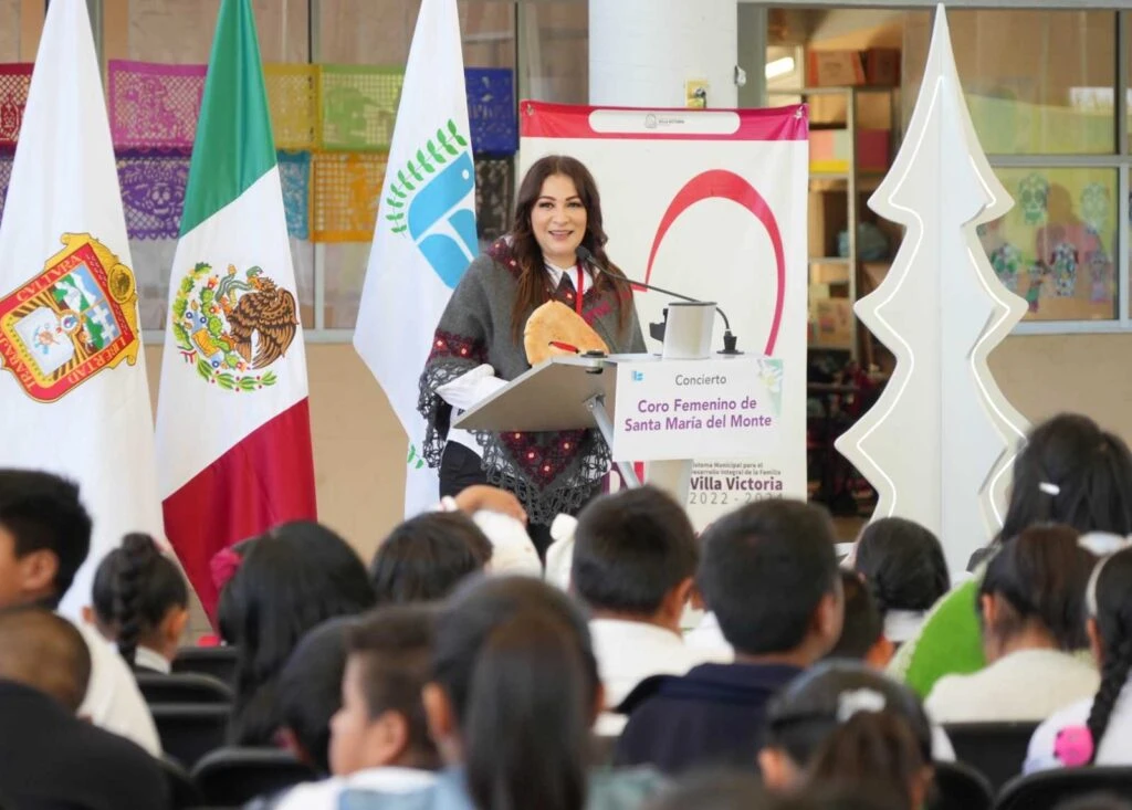 Evento escolar en un espacio techado con gran asistencia de niños sentados en sillas frente a un escenario, donde una persona habla en un atril junto a banderas y elementos decorativos coloridos.