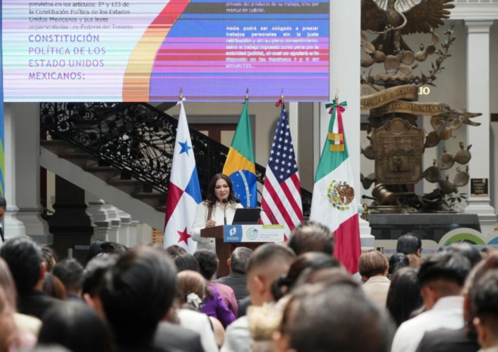 Persona dando un discurso en un podio frente a una multitud, con banderas de distintos países detrás y una pantalla grande que muestra texto sobre la Constitución Política de los Estados Unidos Mexicanos.