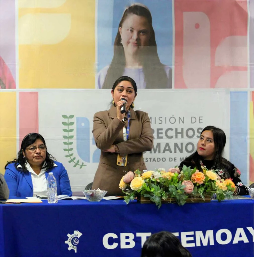 Panel de personas en una mesa con mantel azul durante un evento institucional, donde una persona habla con micrófono frente a un arreglo floral y un fondo con imágenes y el texto de la Comisión de Derechos Humanos del Estado de México.
