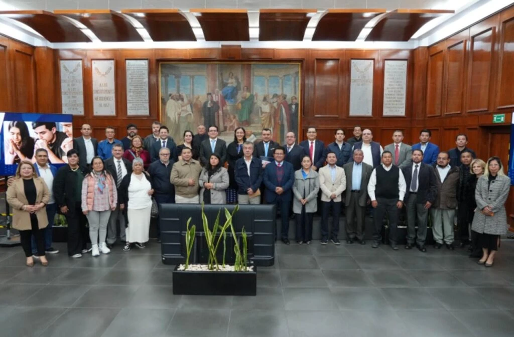 Grupo grande de personas posando en un salón institucional con paredes de madera, frente a un mural histórico y placas conmemorativas, durante una fotografía oficial.