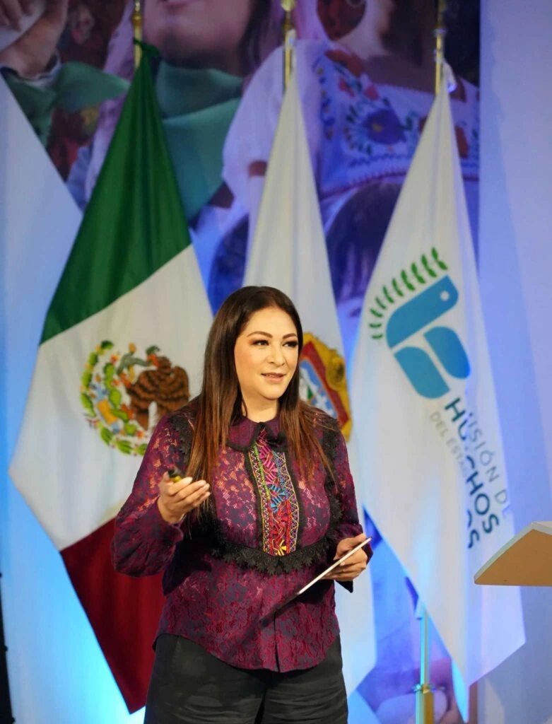 Persona participando en una ponencia frente a banderas oficiales y un fondo institucional, sosteniendo documentos mientras se dirige al público.