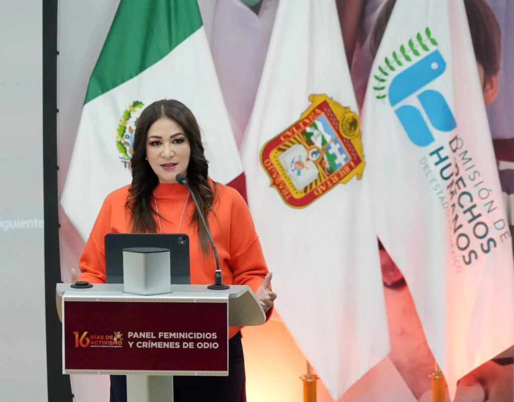Persona exponiendo en un atril durante el evento “Panel Feminicidios y Crímenes de Odio”, con banderas institucionales de fondo y equipo de presentación.
