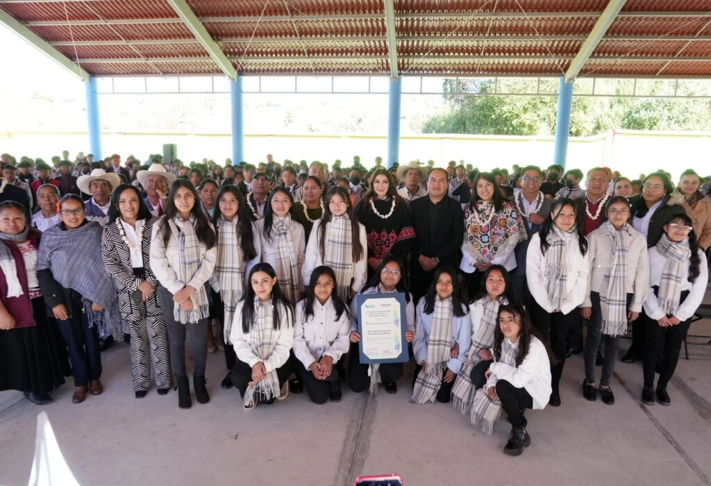 Fotografía grupal de participantes de un coro comunitario y asistentes a un evento cultural, con personas formadas en varias filas sosteniendo un reconocimiento en un espacio techado.
