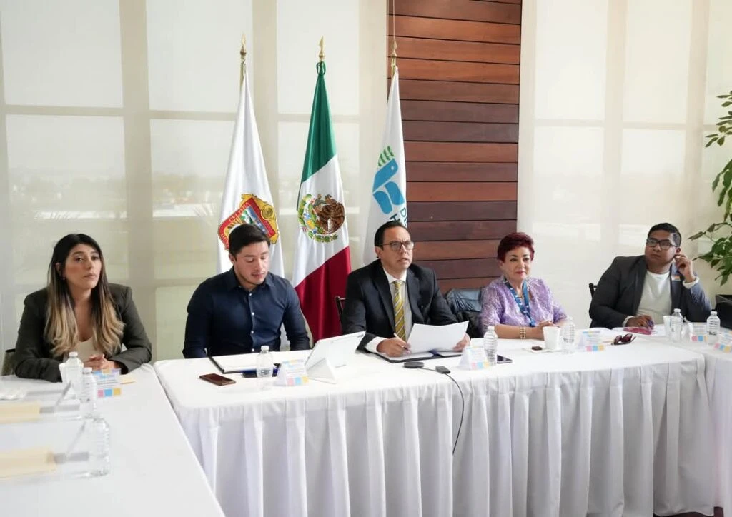 Fotografía de una reunión institucional en la que cinco personas se encuentran sentadas detrás de una mesa larga cubierta con mantel blanco. Sobre la mesa hay botellas de agua, dispositivos electrónicos y documentos