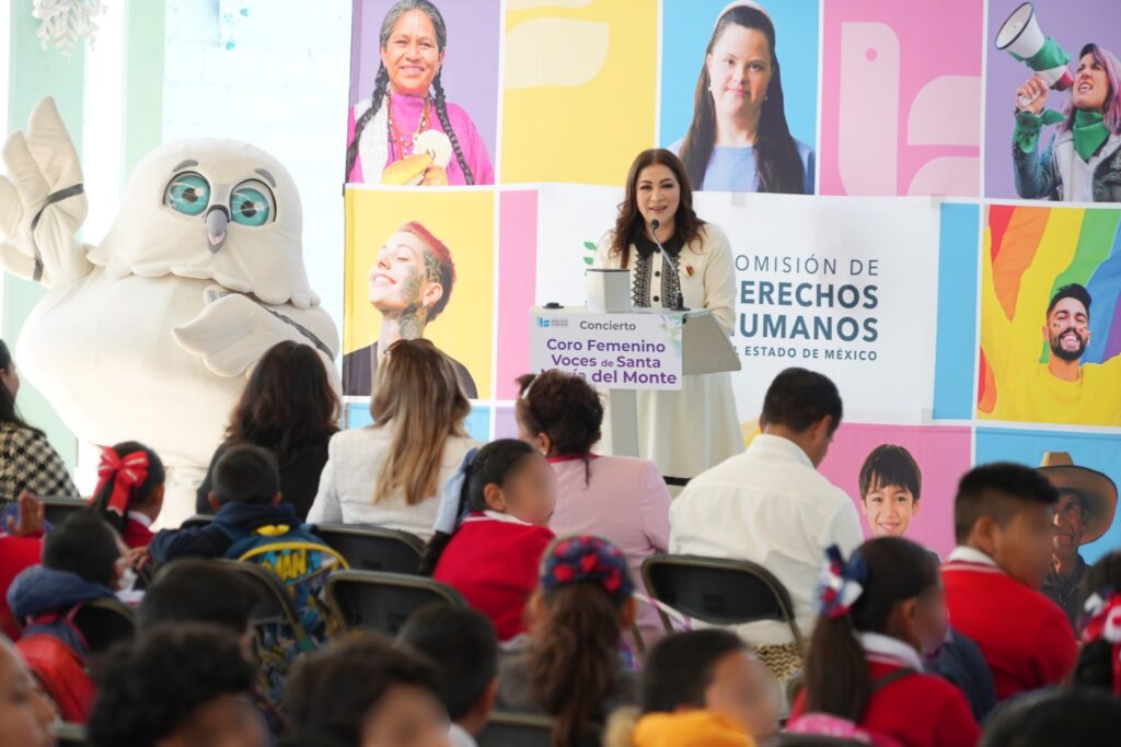 Evento institucional en el que una persona se encuentra en un podio con micrófono frente a un público compuesto principalmente por niñas y niños en uniforme escolar. A un lado hay una mascota institucional con forma de búho. El fondo es un panel colorido con fotografías de personas y el logotipo de la Comisión de Derechos Humanos del Estado de México.