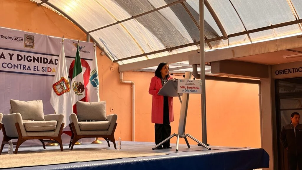 Persona en un podio dando un mensaje durante el panel titulado “VIH, Serofobia y Dignidad”. En el escenario hay sillones, banderas institucionales y un fondo con la temática del evento relacionado con la lucha contra el VIH/SIDA.