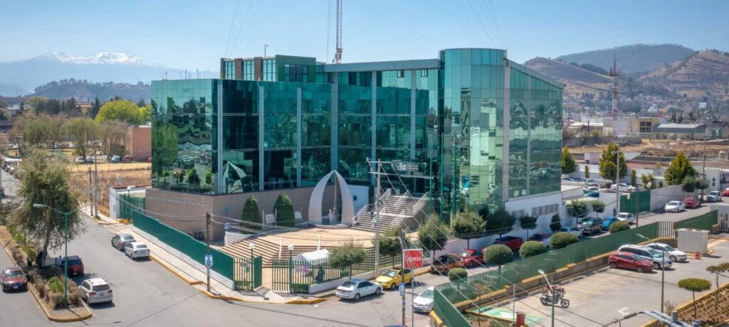 Fotografía de un edificio institucional moderno con fachada de cristal verde ubicado en una esquina urbana. El inmueble está rodeado por una reja perimetral y cuenta con escalinatas de acceso y una escultura en la entrada principal. A su alrededor hay calles con vehículos estacionados y en movimiento. En el fondo se observan montañas, árboles y construcciones urbanas bajo un cielo despejado. Si necesitas el texto en versión más corta o adaptado a accesibilidad web (WCAG), puedo ajustarlo.