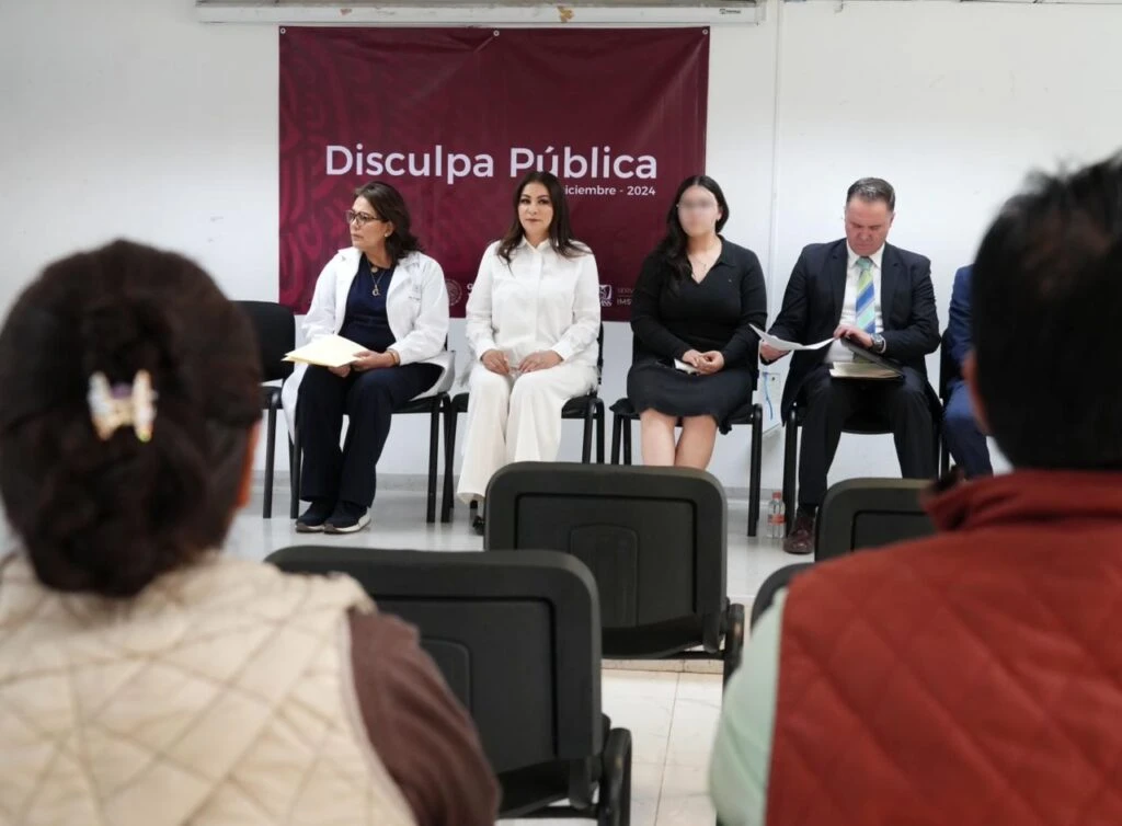 Evento institucional en una sala donde varias personas se encuentran sentadas en una fila frente al público. En la pared posterior hay una lona con el texto “Disculpa Pública” y la fecha de diciembre de 2024. El público está sentado en sillas dispuestas en filas.