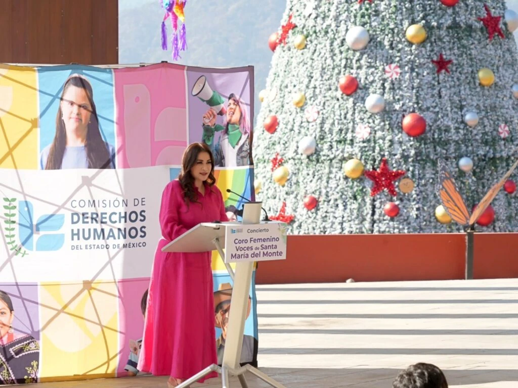 Persona en un podio dando un mensaje durante un evento cultural, junto a un panel con el logotipo de la Comisión de Derechos Humanos del Estado de México. Al fondo se observa un árbol navideño decorado con esferas y figuras, en un entorno al aire libre con paisaje natural.