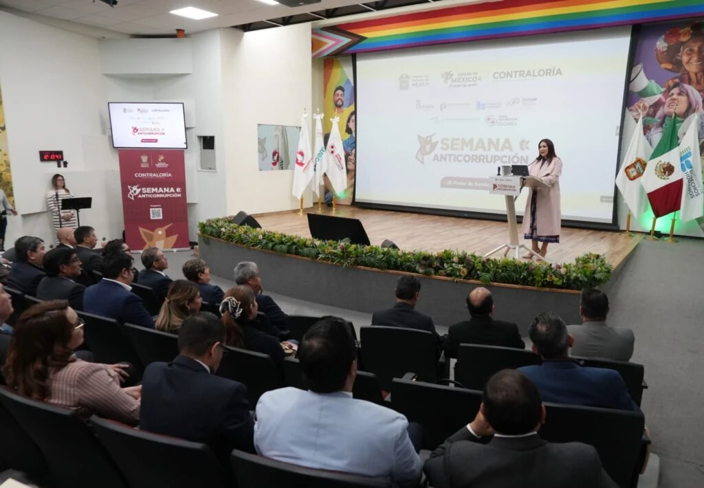 Evento en auditorio con una persona de pie en un podio sobre un escenario de madera, frente a un público sentado en filas. Detrás del podio hay una pantalla grande con el texto “Semana Anticorrupción” y logotipos institucionales, además de banderas colocadas a los lados. La sala cuenta con iluminación uniforme y decoración floral en la parte frontal del escenario.