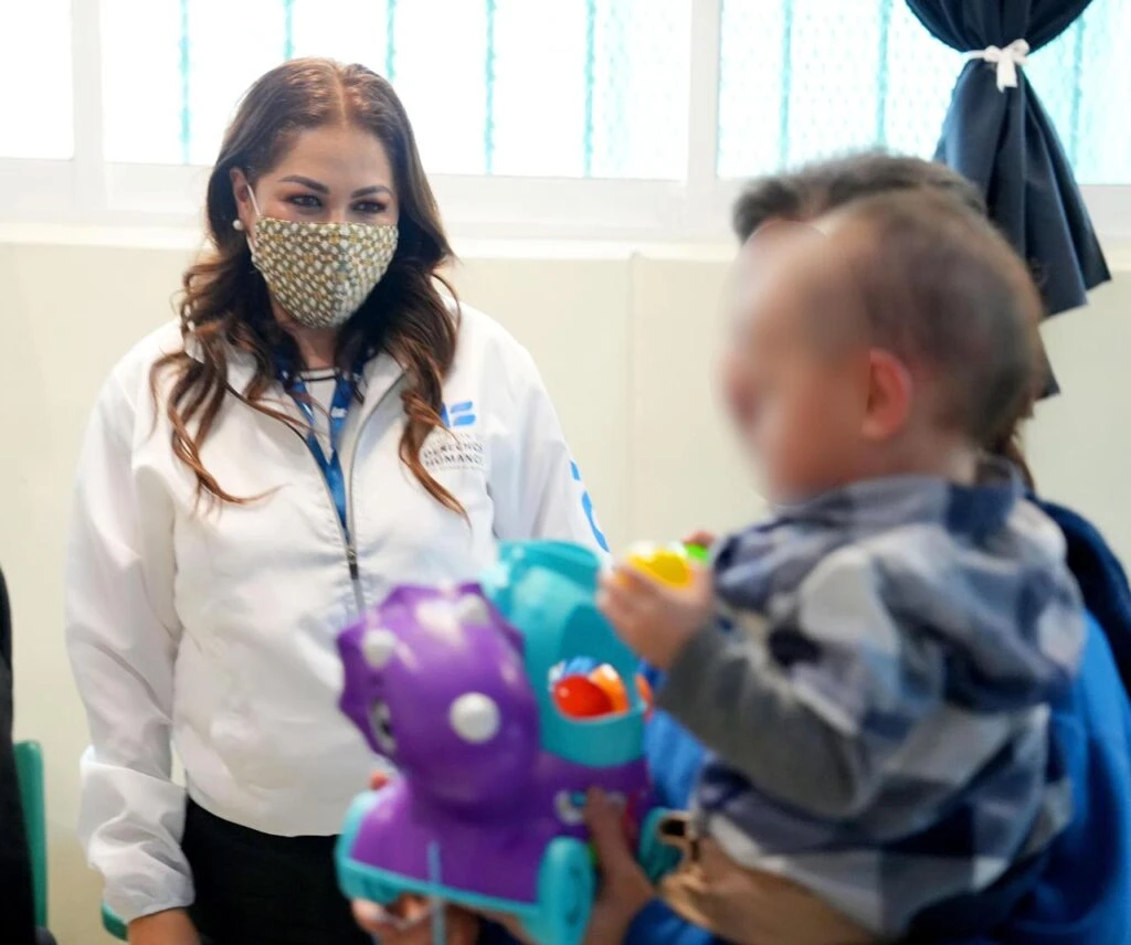Primer plano de una persona con prenda institucional observando a un niño que sostiene un juguete de colores. El fondo muestra una ventana con cortina y paredes claras, en un ambiente interior bien iluminado.