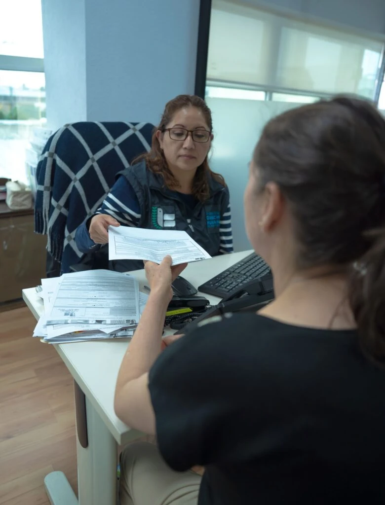 Interior de una oficina donde una persona sentada detrás de un escritorio entrega un documento a otra persona ubicada al frente. Sobre el escritorio hay varios papeles, un teclado y una superficie de trabajo ordenada. El espacio cuenta con iluminación natural proveniente de ventanas laterales y mobiliario de oficina.