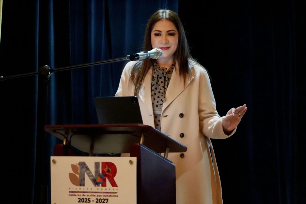 Persona de pie detrás de un atril con micrófono durante una presentación en un auditorio. El atril tiene un logotipo con las letras “NR” y el periodo “2025 - 2027”. Sobre el atril hay una tableta y el fondo es una cortina oscura.