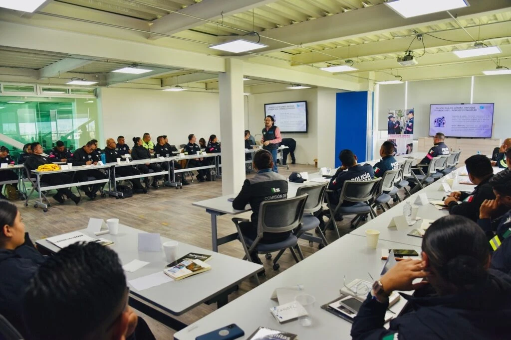 Sala de capacitación con varias personas sentadas alrededor de mesas dispuestas en forma de U. Frente al grupo, una persona está de pie realizando una exposición junto a dos pantallas que muestran información