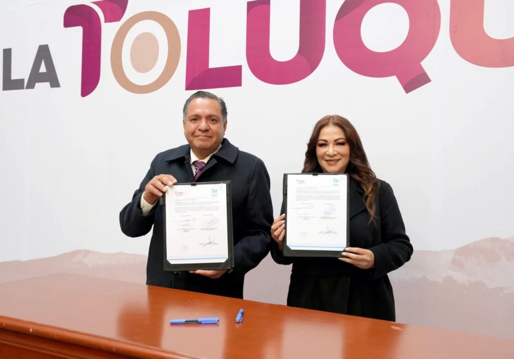 Dos personas de pie frente a una pared con el texto “Toluca” sostienen documentos enmarcados con firmas. En primer plano hay una mesa y bolígrafos, en un espacio de evento institucional con fondo gráfico en tonos claros.
