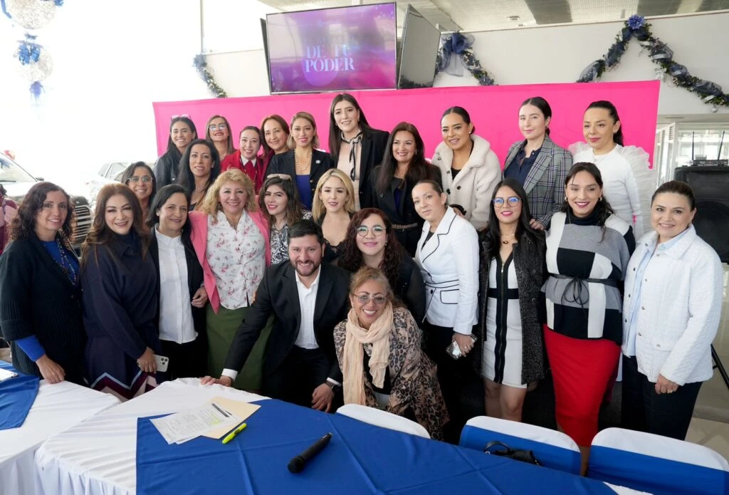 Grupo de personas reunidas en un espacio interior posando para una fotografía alrededor de una mesa con mantel azul. En la parte superior hay una pantalla con el texto “DE PODER” y decoración con guirnaldas. En la mesa hay documentos y un micrófono.