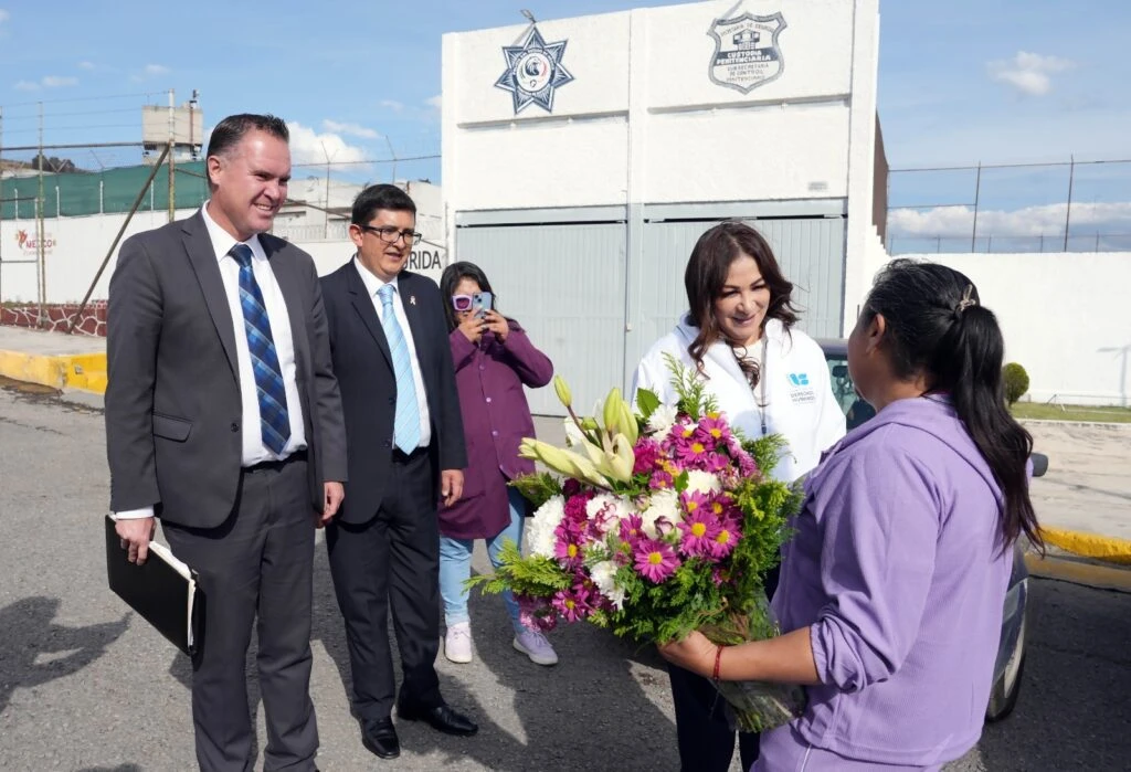 Escena en un espacio exterior junto a una vía cercada. Una persona en primer plano lleva las manos cerca del rostro y sostiene una bolsa transparente, mientras al fondo hay varias personas observando. El entorno incluye una malla metálica, pavimento y edificios cercanos.