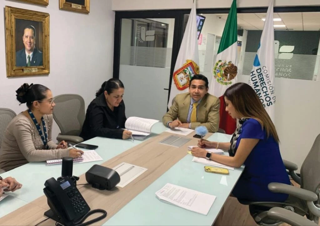 Reunión en una oficina institucional donde cuatro personas están sentadas alrededor de una mesa de trabajo con documentos y dispositivos