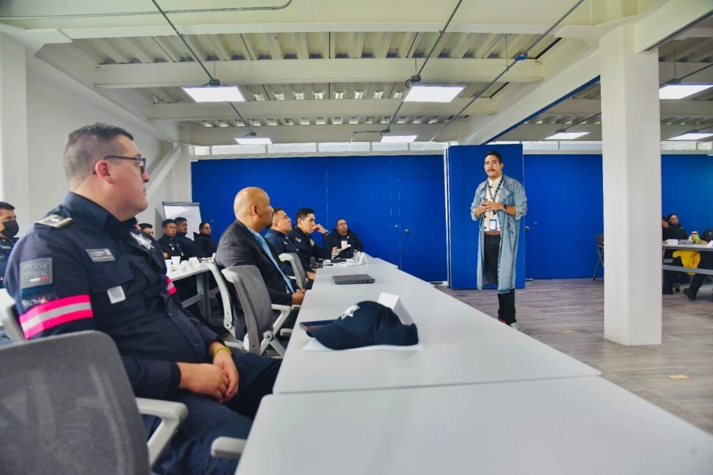 Sala de capacitación con varias personas sentadas alrededor de mesas dispuestas en filas, la mayoría con uniforme oscuro con insignias visibles.