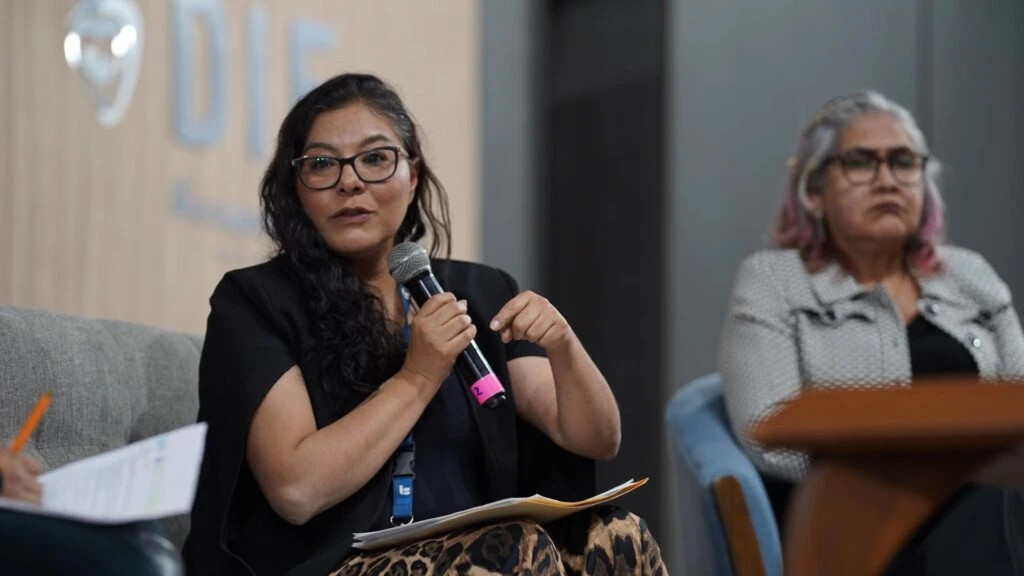 Panel en un evento donde una persona habla sosteniendo un micrófono mientras está sentada en un sillón. A su lado hay otra persona sentada en segundo plano. En primer plano se observan documentos sobre las piernas de la persona que interviene. El fondo incluye un panel institucional con texto parcialmente visible y un entorno interior con iluminación moderada.