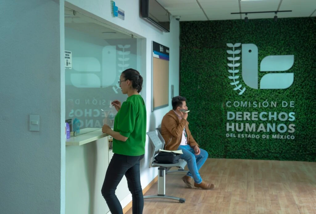Área de atención en una oficina institucional donde una persona se encuentra de pie frente a una ventanilla realizando un trámite. Al fondo, otra persona está sentada en una silla de espera usando un dispositivo móvil