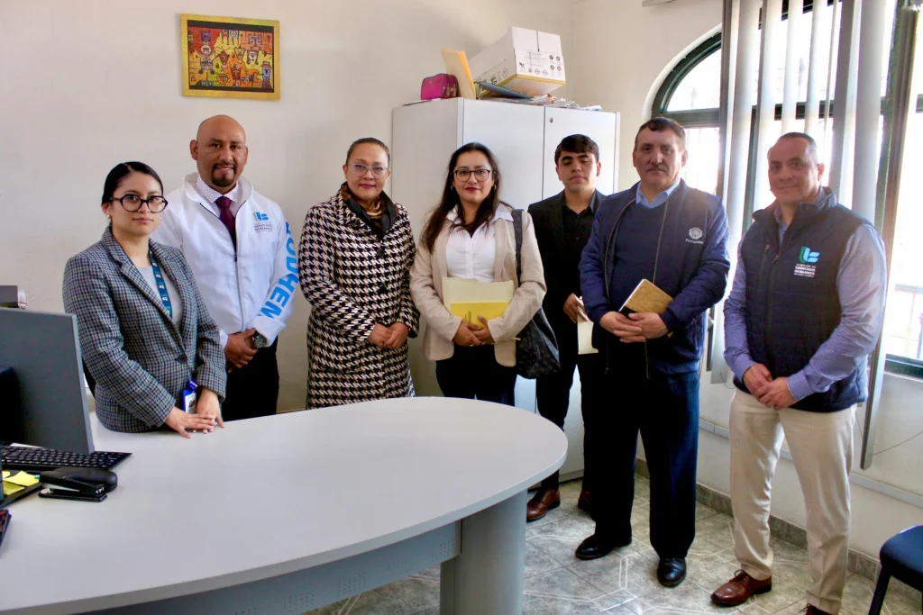Grupo de siete personas posando en una oficina alrededor de un escritorio. Algunas personas sostienen carpetas o documentos y llevan identificaciones visibles. En el fondo hay un archivero con cajas encima, una ventana con persianas y un cuadro colorido colgado en la pared. El espacio cuenta con iluminación natural y mobiliario de oficina.
