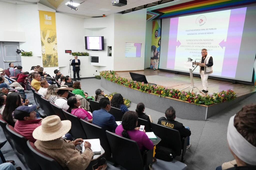 Auditorio con público sentado en butacas observando una presentación en un escenario. Una persona se encuentra en un podio frente a una pantalla grande con texto y gráficos. El escenario está decorado con arreglos florales y elementos visuales en colores vivos. En las paredes hay murales y pantallas adicionales.