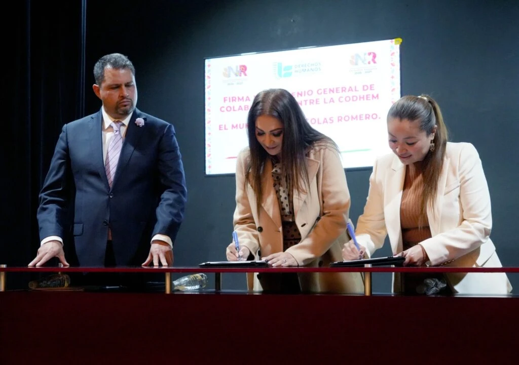 Escenario interior donde dos personas firman documentos sobre una mesa durante un evento. A un lado, otra persona observa de pie. En el fondo se proyecta una pantalla con texto que incluye “Firma convenio general de colaboración entre la CODHEM…”. El espacio cuenta con iluminación focal sobre el escenario y fondo oscuro.