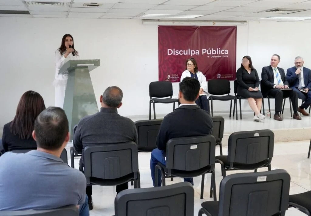 Evento en una sala donde una persona se encuentra de pie hablando desde un podio con micrófono. Detrás hay una lona con el texto visible “Disculpa Pública” y varias personas sentadas en el escenario en sillas. En el área frontal se observan asistentes sentados en filas de sillas de plástico, en un espacio interior con iluminación uniforme.