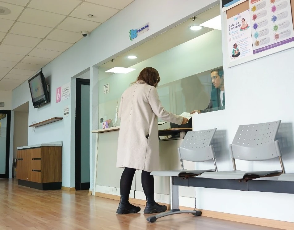Interior de un área de atención institucional donde una persona se encuentra de pie frente a una ventanilla entregando documentos a otra persona ubicada detrás de un vidrio. El espacio incluye sillas metálicas de espera, carteles informativos en la pared, un monitor en la parte superior y mobiliario en tonos claros con iluminación artificial.