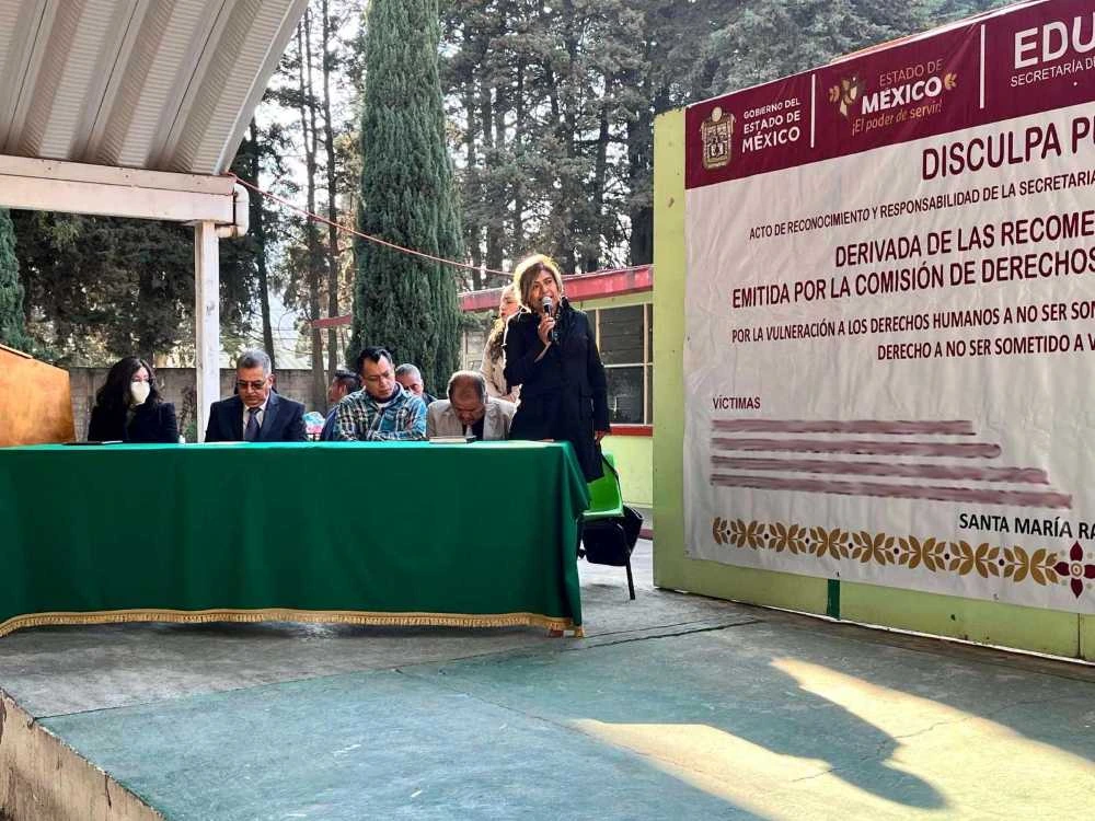 Vista amplia de un evento bajo una estructura techada, con asistentes sentados frente a una mesa principal y un panel grande con texto institucional al lado izquierdo, en un entorno escolar o comunitario.