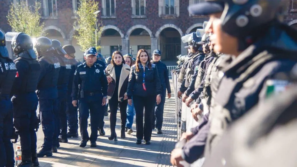Recorrido oficial entre dos filas de policías estatales, donde varias personas caminan al centro en un espacio abierto con edificaciones al fondo.