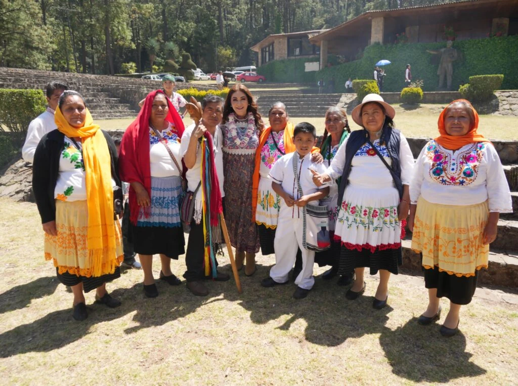 Grupo de personas en exterior con vestimenta tradicional colorida posando frente a una construcción rodeada de árboles; algunas personas usan prendas bordadas y accesorios típicos, formando una fila frente a la cámara.