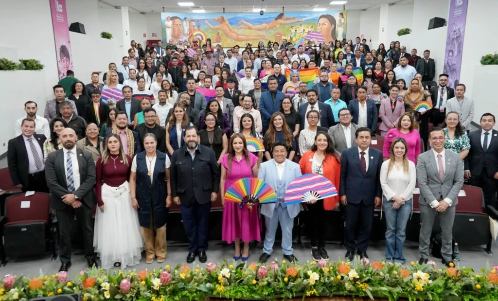 Persona en un podio dando un mensaje formal en un escenario, con banderas institucionales detrás y un cartel que hace referencia a la firma de convenios con poblaciones LGBTTTIQA+ mexiquenses.