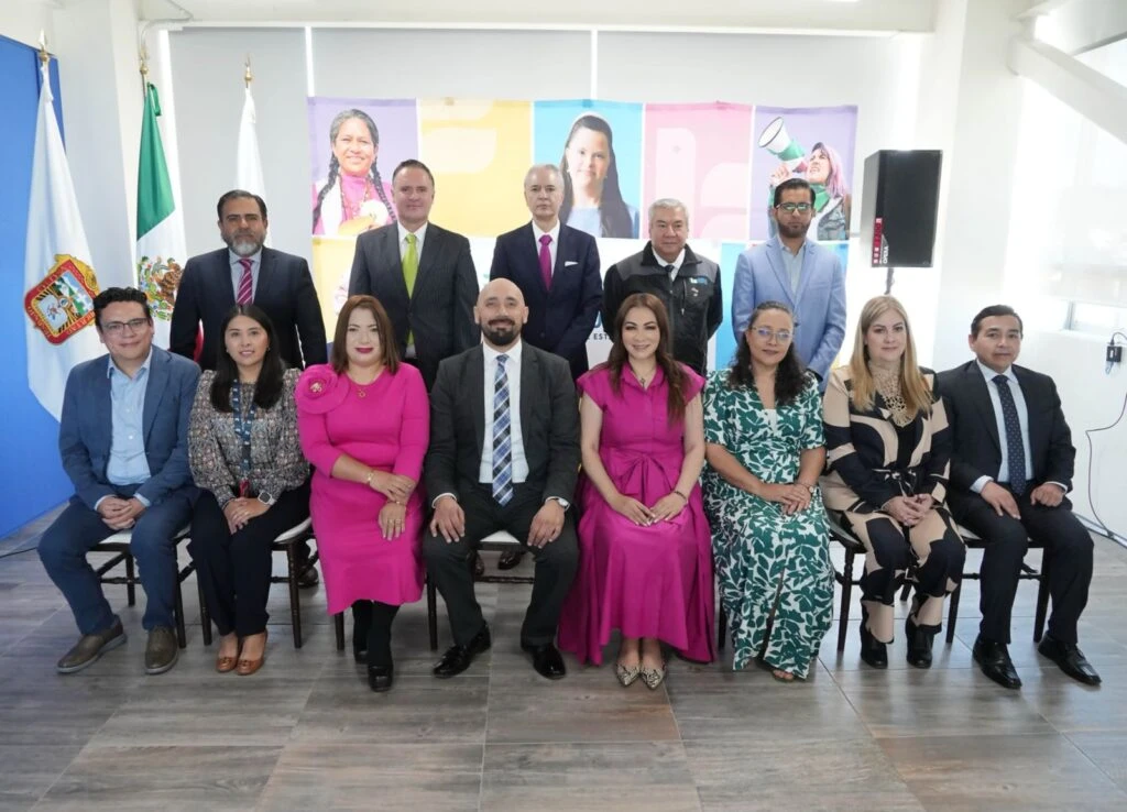 Grupo de personas posando en dos filas en un salón institucional, con banderas a un lado y un mural colorido al fondo, durante un evento oficial.