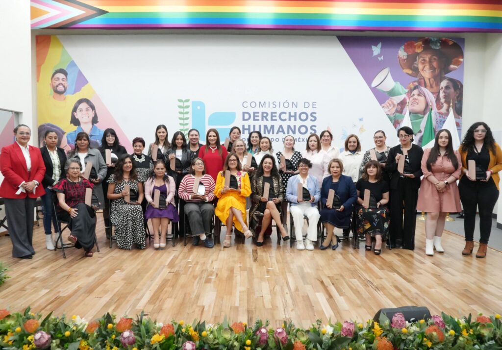 Personas en una ceremonia sostienen un reconocimiento y una placa, acompañadas por otras participantes que aplauden frente a un fondo institucional de derechos humanos.