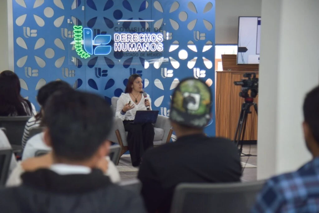 Escena de conferencia con una persona hablando desde un asiento frente a público asistente, con equipo de grabación y un fondo institucional con logotipos de derechos humanos.