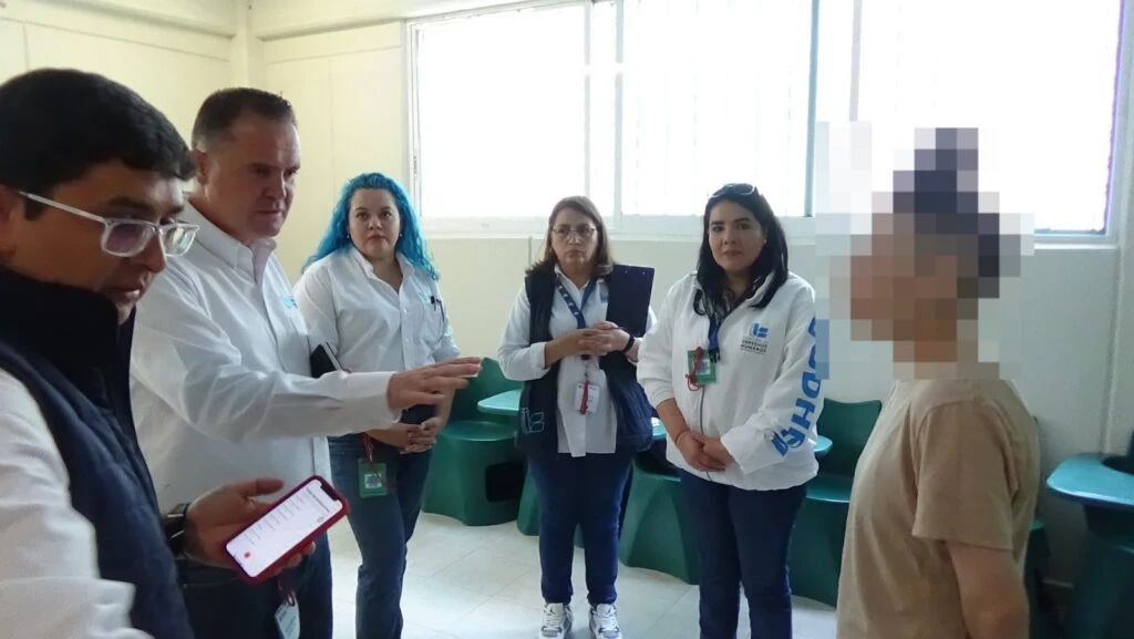 Grupo de personal institucional de pie durante una visita de supervisión en un espacio interior, dialogando con una persona cuya identidad está protegida, en una sala con mobiliario fijo.