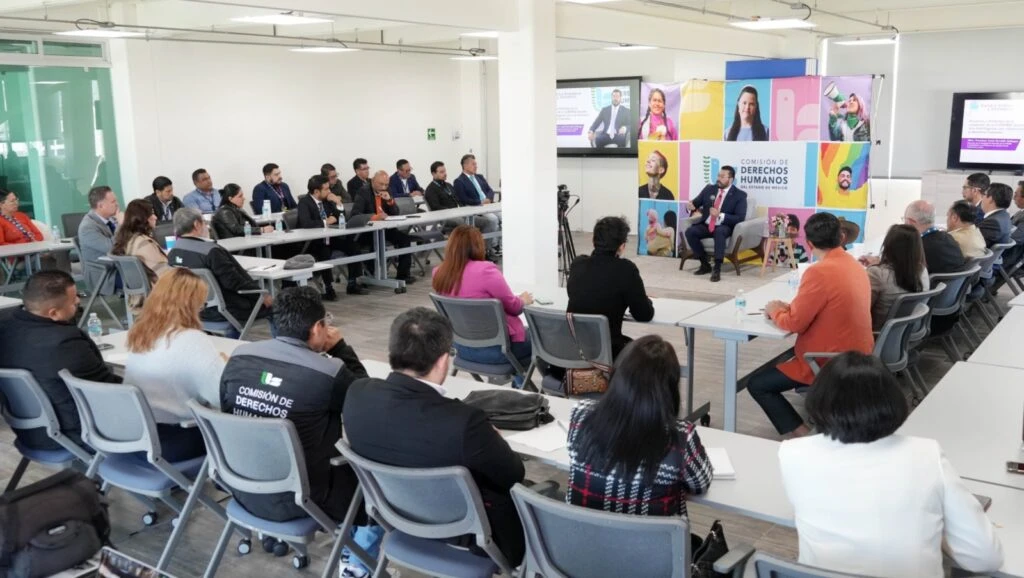 Sala de conferencias con múltiples asistentes sentados en mesas mientras una persona expone al frente, con material institucional y un fondo gráfico colorido de la Comisión de Derechos Humanos.