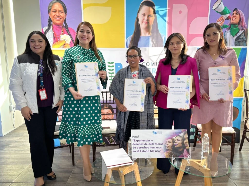 Grupo de personas posa mostrando reconocimientos impresos durante un evento institucional, frente a un fondo con imágenes diversas y material gráfico relacionado con derechos humanos.