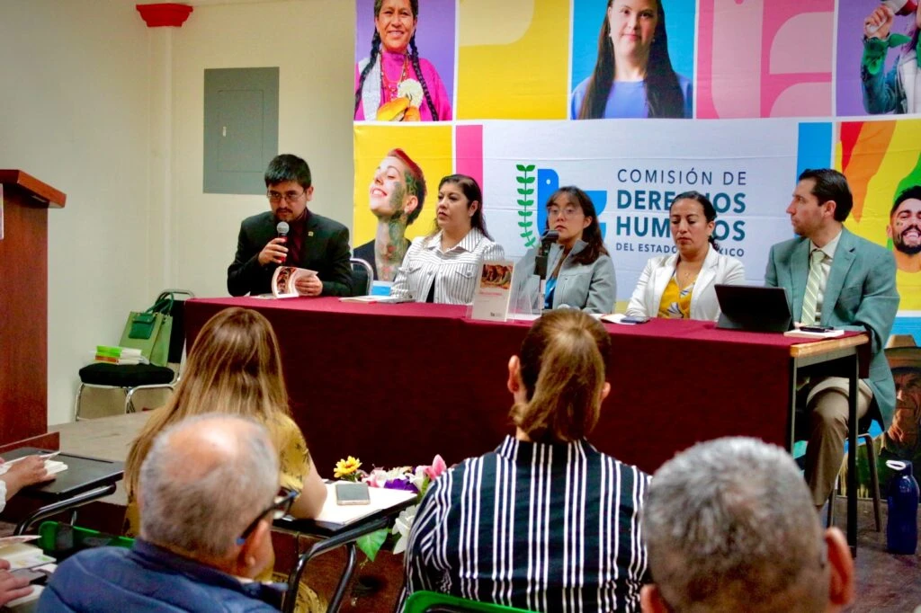 Panel de personas sentadas detrás de una mesa durante una presentación editorial, frente a un público asistente, en un salón con fondo institucional de la Comisión de Derechos Humanos del Estado de México.