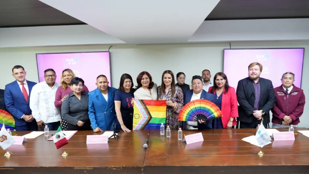 Grupo numeroso de personas posando en un salón institucional detrás de una mesa, algunas sosteniendo banderas arcoíris y abanicos multicolores, con pantallas y señalización al fondo.