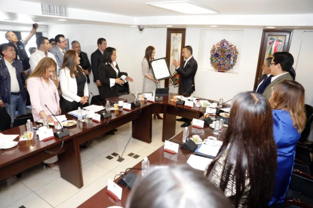 Reunión institucional en sala de juntas con varias personas alrededor de una mesa en forma de U, mientras se entrega un documento enmarcado a una persona al centro, con asistentes de pie y sentados observando el acto.