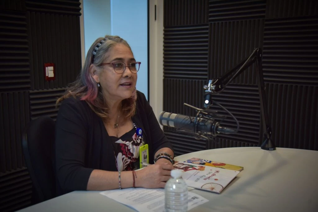 Persona sentada en un estudio de grabación con paneles acústicos en las paredes, hablando frente a un micrófono profesional montado en brazo articulado, con documentos y un libro sobre la mesa junto a una botella de agua.