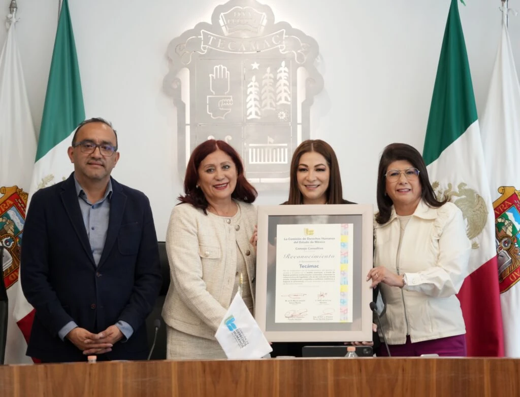 Grupo de varias personas posando en una sala institucional mientras sostienen un reconocimiento enmarcado al centro, con banderas y un escudo oficial en la pared de fondo.