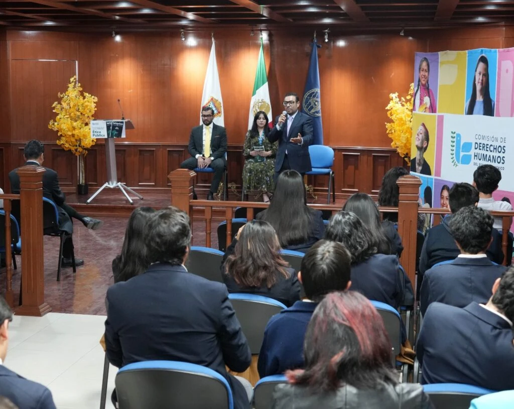 Vista desde el público de una charla en un auditorio pequeño, donde una persona expone al frente acompañada por otra persona sentada, con banderas institucionales y un cartel lateral de derechos humanos.